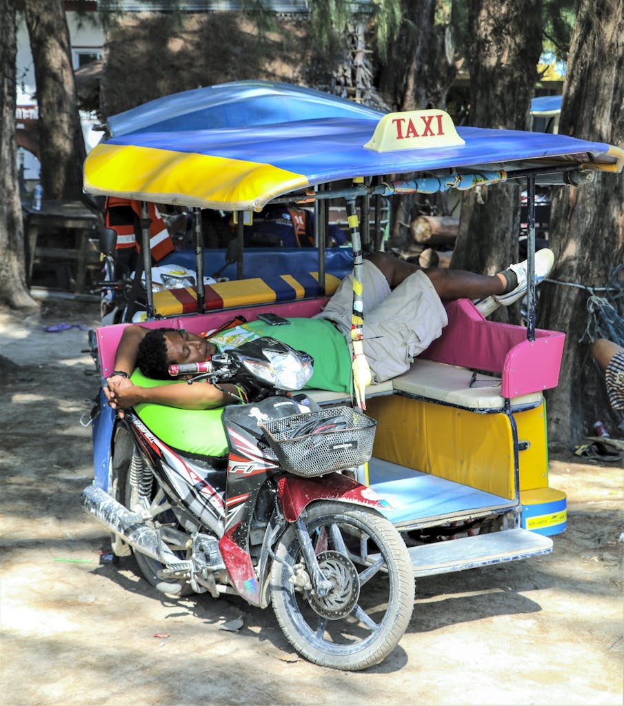 Asian man sleeping on a colorful motorbike tricycle taxi under trees in Thailand.